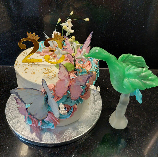 23ste verjaardagstaart gedecoreerd met roze en blauwe bloemen met daarop vlinders in wit en roze. Bovenop de taart staan gouden getallen die samen het getal 23 vormen. Naast de taart staat een groene vogel gemaakt van suiker.