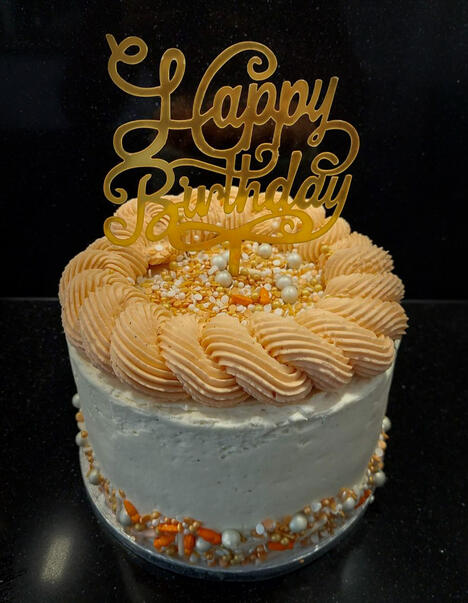 Witte taart gedecoreerd met een gouden rand. Het midden van de taart is gevuld met gouden en witte balletjes. Bovenop de taart staat de tekst 'Happy birthday' in gouden letters.